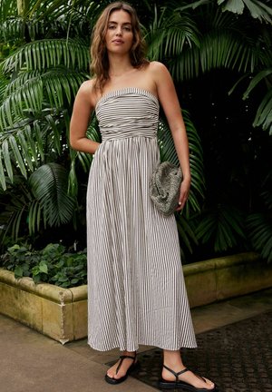 Gestreiftes, trägerloses Maxikleid aus schwarz-weißem Stoff mit gerafftem Oberteil, das einen fließenden Rock und eine Seitentasche enthält. Getragen mit schwarzen Sandalen.