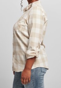 Persona che indossa una camicia a maniche lunghe beige e bianca a scacchi con le maniche arrotolate e jeans azzurri chiari, in piedi di lato su uno sfondo semplice.