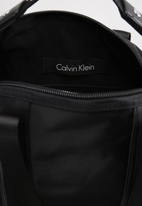 Zwarte tas met een gladde textuur, voorzien van een ritssluiting en een binnenlabel waarop "Calvin Klein" staat op een stoffen label.