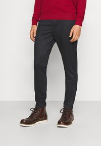 Pantalons taillés gris foncé avec un motif texturé, associés à des bottines en cuir marron avec des semelles et des lacets blancs. Un pull rouge au-dessus.