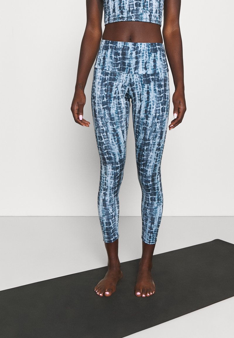 Femme se tenant pieds nus sur un tapis de yoga noir, portant un legging court à motifs bleus et un soutien-gorge de sport assorti, devant un fond uni.