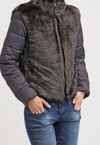 Szary kamizelka z futra syntetycznego z wysokim kołnierzem, w połączeniu z pikowanymi szarymi rękawami i zamkiem z przodu, noszona na niebieskich dżinsach.