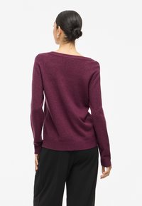 Burgundijas krāsas adīts džemperis ar plašu kakla izgriezumu, garām piedurknēm un ribinātu apakšmalu, kas izceļ mīkstu tekstūru un brīvu piegriezumu. Zem tā redzami melni bikses.