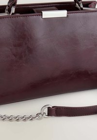 Sac à main en simili cuir bordeaux avec une texture lisse, quincaillerie argentée et une bandoulière en chaîne. Il présente une forme rectangulaire plate et un design simple.
