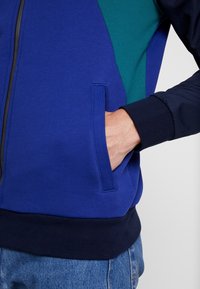 Modrá a zelená barevně blokovaná mikina s kapucí na zip, která se vyznačuje texturovanou tkaninou, boční kapsou, pružnými manžetami a kontrastním námořnickým páskem.