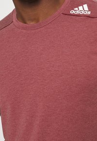 Bordový sportovní tričko vyrobené z měkké, strukturované tkaniny. Obsahuje kulatý výstřih a bílé logo Adidas na levém rameni.