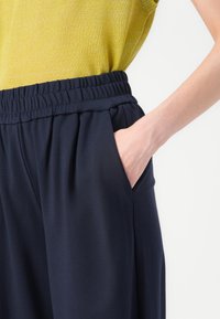 Main dans la poche d’un pantalon ample bleu foncé avec ceinture élastique, associé à un top en maille jaune sans manches sur fond clair.