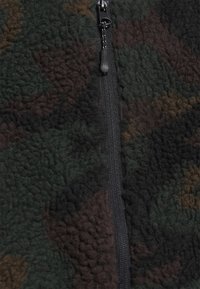 Närbild av en svart dragkedja på mörkgrön, brun och svart kamouflagemönstrad fleece.