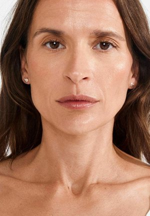 Vrouw met middellang bruin haar, lichte huid en subtiele make-up; draagt oorbellen in de vorm van een hart; neutrale gezichtsuitdrukking, blote schouders.