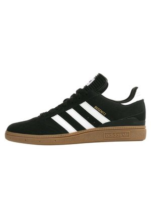 Zapatillas de ante negro con tres franjas blancas, suela de goma y un cuello acolchado. Cuenta con letras doradas "BUSENITZ" en el costado.