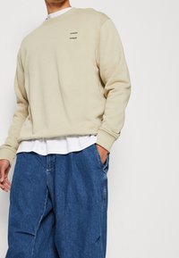 Béžová bavlněná mikina s žebrovaným kulatým výstřihem a malým černým logem značky. Nositá přes bílou vrstvu s volnými modrými denimovými kalhotami.