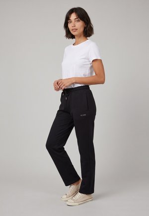 Femme debout portant un t-shirt blanc à manches courtes, un pantalon noir à cordon avec le logo GUESS, et des baskets basses beiges sur un fond gris uni.