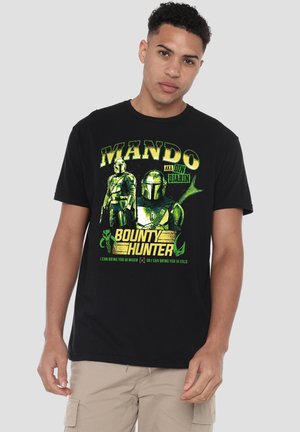 Junger Mann trägt ein schwarzes T-Shirt mit grün-gelber Grafik eines Mandalorian-Helms und Rüstung, Text "Mando AKA Din Djarin Kopfgeldjäger."