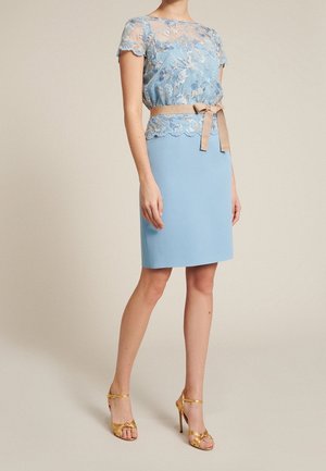 Femme portant un haut en dentelle bleu clair avec broderie florale, une jupe assortie longueur genou, une ceinture ruban beige et des sandales à talons hauts dorées.