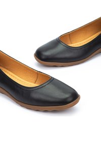 Ballerines noires en cuir avec un bout rond, intérieur beige et semelle en caoutchouc flexible. Texture lisse avec un design minimaliste et sans matériel visible.