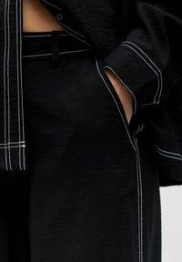 Gros plan sur une personne portant un pantalon et une chemise noirs avec des surpiqûres blanches en contraste, main glissée dans la poche, mettant en valeur la texture du tissu et les détails du design.