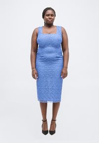 Γυναίκα με κοτσιδάκια, φορώντας μπλε δαντελωτό φόρεμα χωρίς μανίκια μέχρι το γόνατο και μαύρα τακούνια με λουράκι στον αστράγαλο, στέκεται μπροστά σε ένα απλό λευκό φόντο.