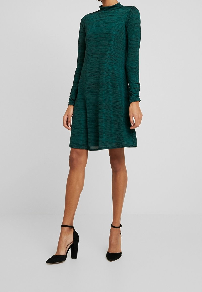 Femme portant une robe vert foncé à manches longues, longueur genoux, avec des chaussures noires à talons hauts et à bride cheville, debout devant un fond uni.