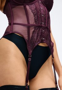 Burgunderfarbene Spitzencorsage mit durchscheinenden Mesh-Einsätzen, verstellbaren Straps und silberfarbenen Beschlägen. Kombiniert mit einem dunklen, hochgeschnittenen schwarzen Slips.
