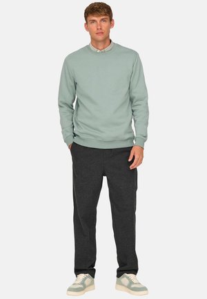 Nuori mies seisoo päällään vaaleanvihreä collegepaita harmaan kauluspaidan päällä, tummanharmaat housut ja vaaleanvihreät tennarit, kädet taskuissa.