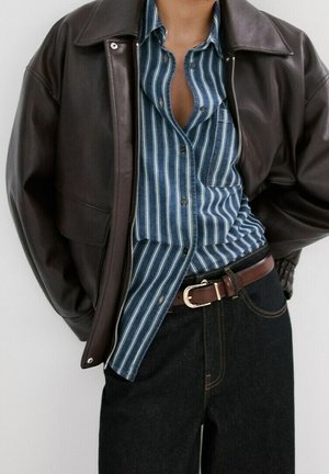 Personne portant une veste en cuir brun foncé ouverte sur une chemise rayée verticalement bleue et blanche rentrée dans un jean foncé avec une ceinture marron.