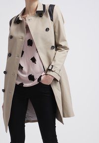 Trench coat beige con accenti neri sulle spalle, bottoni doppi, e vita fissata con una cintura. Indossato su una maglietta rosa con macchie nere.