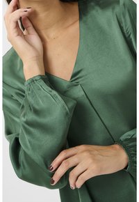 Blouse en satin vert avec un col en V, des manches bouffantes à poignets élastiques, et une texture lisse et brillante.