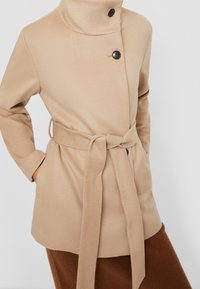 Beige jas van zachte stof met een hoge kraag, voorzien van een ceintuur, zijzakken en twee zwarte knopen om te sluiten.