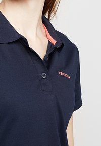 Mörkblå polo med en tvåknäppt krage, med en korallfärgad accent på kragen och en liten logotyp på vänster bröst. Slät textur.