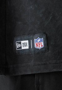 Schwarzer Stoff mit glatter Textur zeigt ein Etikett mit den Logos von "New Era" und "NFL" in Weiß und Rot auf weißem Hintergrund.