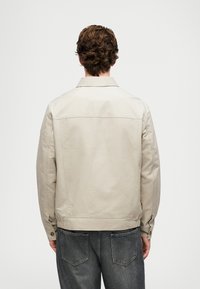 Vyras, užsidėjęs šviesiai kreminę striukę ir tamsiai pilkas džinsus, fotografuotas iš už nugaros prieš baltą fono.