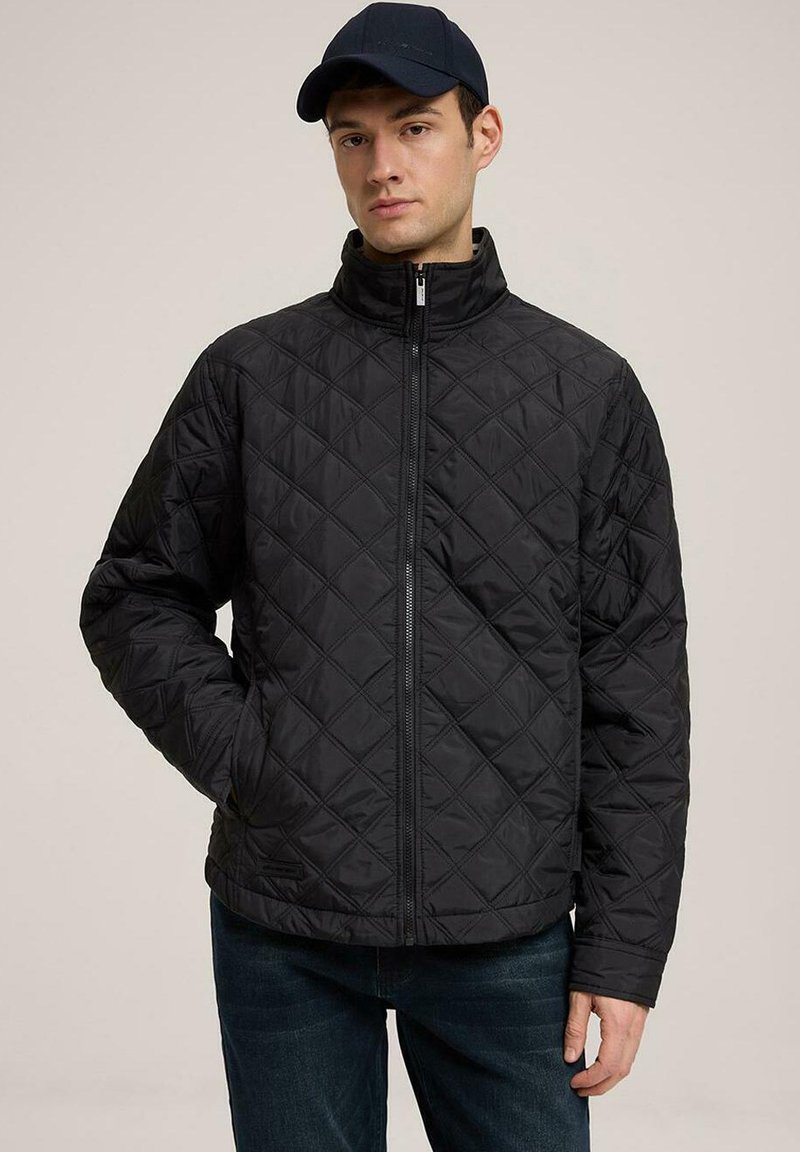 Czarna pikowana kurtka z wysokim kołnierzem, pełnym zamkiem z przodu i bocznymi kieszeniami. Materiał ma teksturowany wzór w diamenty, zestawiony z ciemnoniebieskimi dżinsami.