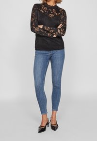 Sort blonde langærmet top med blomstermønster, høj hals, parret med blå skinny jeans og sorte spidse hæle.