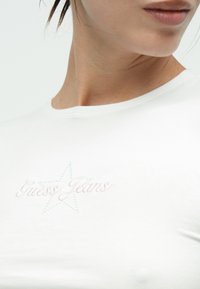 Λευκό βαμβακερό t-shirt με πολύχρωμο κεντητό κείμενο "Guess Jeans" και σχέδιο αστεριού. Απλός στρογγυλός λαιμός με λεία υφή.