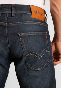 Mörk denimjeans med orange sömmar, föreskrivna med en läderetikett där det står "REPLAY BLUEJEANS" och en vågmönster på bakfickorna.