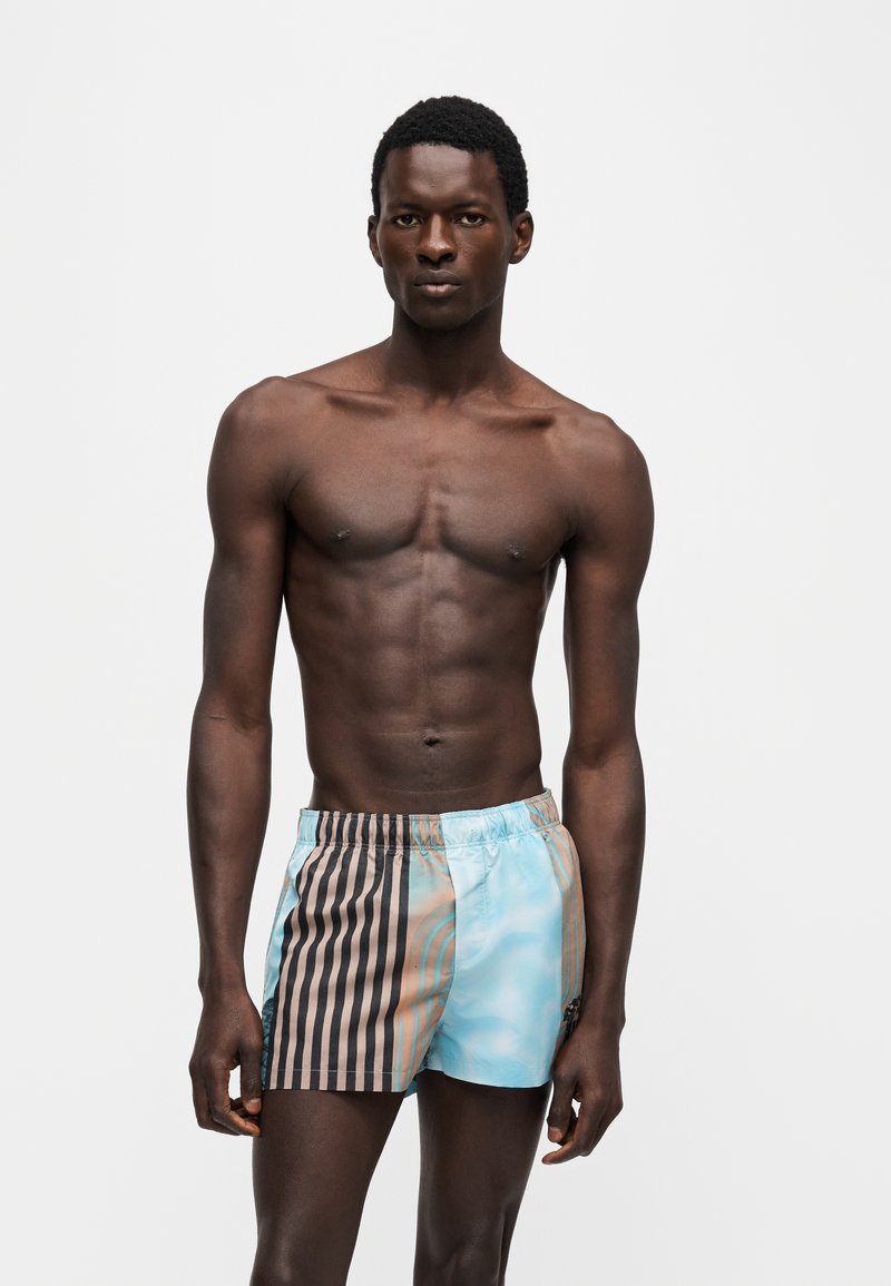 Homme torse nu debout face à l’avant, portant un short de bain rayé bleu pastel et marron sur fond blanc uni.