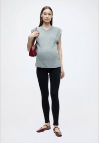 Sivý rebrovaný tielko bez rukávov so zapínaním na bočných gombíkoch, v kombinácii s čiernymi legínmi a bordovými balerínami, držený s malou červenou kabelkou.