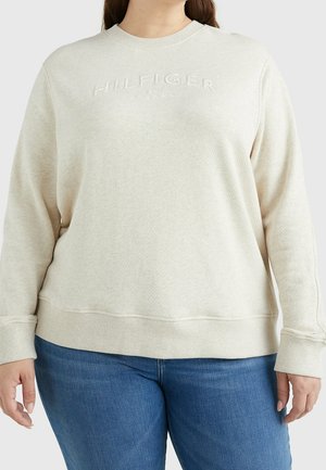 Persona con sudadera de color beige claro con "HILFIGER" bordado en el pecho, combinada con vaqueros azules, de pie frente a un fondo liso.