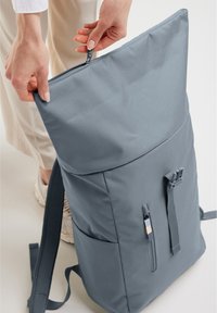 Zaino in tessuto grigio con chiusura roll-top, tasca frontale con zip, spallacci regolabili e superficie testurizzata. Presenta un design minimalista.