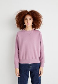 Lys lilla hettegenser med ribbestrikket kant og mansjetter, med rund hals og en sydd logo detalj nær kanten. Brukt sammen med mørke jeans.