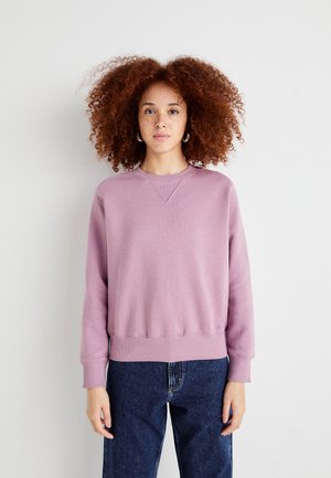 Light purple sweatshirt with a ribbed hem and cuffs, featuring a crew neck and a stitched logo detail near the hem. Worn with dark jeans.