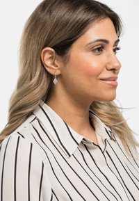 Orecchini a cerchio in argento con un design a forma di diamante e dettagli testurizzati, indossati da un modello con capelli lunghi e ondulati, vestito con una blusa a righe.