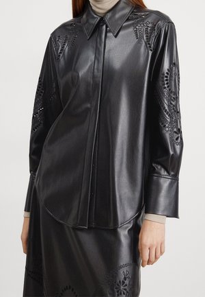 Zwarte leren blouse met lange mouwen en bloemige uitsnijdingen op de mouwen en schouders, gedragen over een beige coltrui.