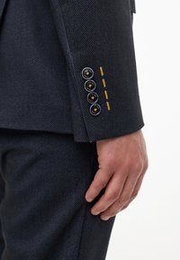 Blazer in lana con motivo a spina di pesce di colore blu scuro. Caratterizzato da quattro bottoni sui polsini e dettagli di cucitura gialla. Vestibilità strutturata.
