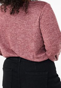 Persona che indossa un maglione rosa polveroso texturizzato infilato in jeans neri a vita alta con tasche posteriori, capelli ricci visibili all'altezza del colletto.