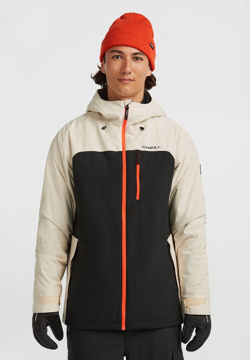 Chaqueta aislante para hombre con la parte superior beige y la parte inferior negra, detalles en naranja brillante, bolsillos con cremallera y capucha. Se llevan guantes en las manos.