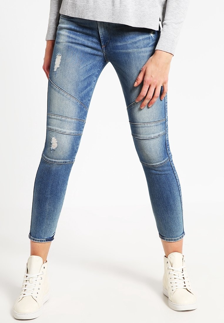 Vaqueros de mezclilla azul claro con parches desgastados y detalles de costura horizontal. Presentan un corte slim fit y longitud hasta los tobillos, combinados con zapatillas blancas.