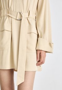 Robe beige avec une taille ceinturée, des manches longues et deux poches avant. Texture douce et design épuré. Quincaillerie en argent sur la ceinture.