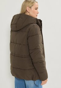 Brun puffjacka med huva, med quiltat mönster och synlig sömnad. Texturerad tyg och dragsko vid fållen.