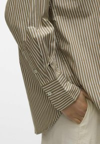 Brun og hvitstripete skjorte med lange ermer, knappede mansjetter, og myk stofftekstur, delvis stappet inn i lyse bukser.